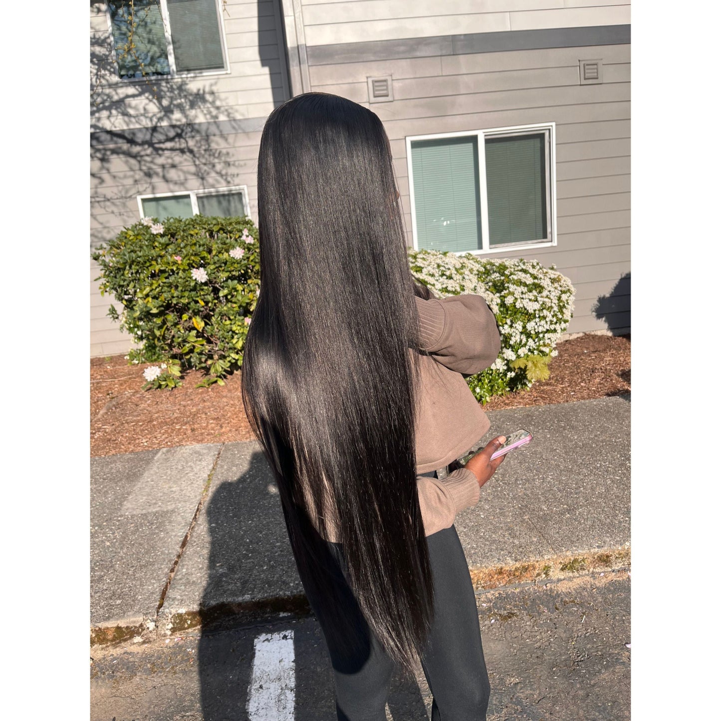 Person with long black raw hair bundles standing on a sidewalk with a house and garden in the background