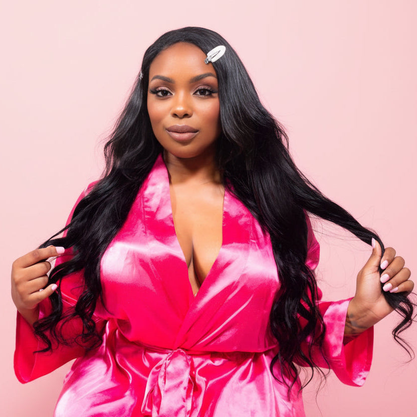 Woman in a pink satin robe holding black hair extensions against a pink background
